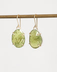 A pair of gold drop earrings with organic-shaped light green gemstones, hanging from a gold-colored horizontal bar against a plain white background.