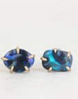 A pair of stud earrings featuring irregular, polished blue opal stones with gold prong settings, displayed on a clean white background.