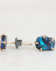 A pair of gold stud earrings featuring rectangular blue opal gemstones, with one earring shown from the side/back and the other from the front, resting on a light gray surface.