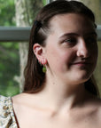 A close-up portrait of a woman wearing a small green teardrop dangle earring and a matching green hoop earring, photographed in natural light in front of a window.