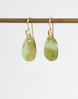 A pair of gold-tone drop earrings with polished light green teardrop-shaped stone pendants, hanging from a horizontal gold rod against a plain white background.