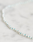 A delicate beaded necklace with white pearl-like beads and small turquoise blue accent beads, photographed on a light marble surface.