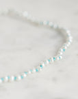 A close-up of a delicate beaded strand necklace with white pearl-like beads and small turquoise blue accents, resting on a light marble surface.