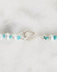A close-up of a beaded bracelet featuring white pearl-like beads, turquoise blue knotted accents, and a textured silver clasp, displayed on a light marble surface.