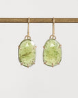 A pair of oval green gemstone drop earrings in gold-toned prong settings hanging from gold-toned hooks, suspended from a horizontal gold rod against a light gray background.