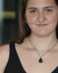 A woman wearing a dark green teardrop pendant necklace on a black chain, paired with matching dark blue teardrop earrings, shown in a close-up portrait.