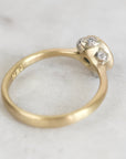 A close-up of a vintage yellow gold ring with a textured, organic-shaped setting holding three small clear stones, photographed on a light gray marble surface.