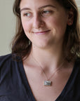 A woman wearing a silver-toned rectangular pendant necklace with a floral engraving, paired with a dark navy blue v-neck t-shirt.