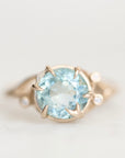 A gold ring featuring a large round-cut light blue gemstone in a prong setting, with small accent stones on the band, photographed against a soft white background.