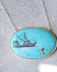 An oval turquoise pendant necklace featuring a hand-painted black and white ship illustration, set in a gold-tone frame on a silver chain, displayed on a textured gray stone surface.