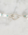 A close-up of a pearl necklace with a silver fishhook clasp and turquoise cord accents, laid on a light marble surface.