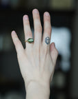 A hand wearing a gold ring with an oval green stone, shown against a dark, blurred background. The hand also features a small geometric tattoo on one of the fingers.