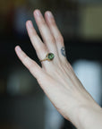 A gold ring with a round green gemstone worn on a person's finger, shown against a dark, blurred background.