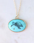 An oval pendant necklace featuring a turquoise blue background with a black ink-style illustration of a sea turtle, set in a gold-tone bezel and hanging from a gold chain on a light marble surface.