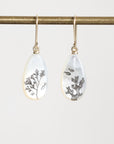 A pair of gold-tone drop earrings with teardrop-shaped white mother-of-pearl pendants featuring black botanical and fish illustrations, hanging from a gold rod against a light gray background.