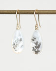 A pair of teardrop-shaped white earrings with gold fishhook wires, hanging from a gold rod. One earring contains dried botanical elements, and the other features a small fish and coral design.
