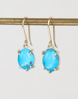 A pair of gold drop earrings with oval turquoise-blue gemstones in a prong setting, hanging from a gold rod against a light gray background.