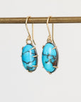A pair of gold drop earrings with oval turquoise-blue marbled stones, hanging from a gold rod against a light gray background.