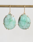 A pair of gold drop earrings with irregular light green gemstone pendants, hanging from a gold bar against a light gray background.