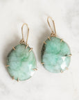 A pair of gold drop earrings with faceted light green gemstone pendants, displayed on a light marble surface.