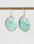 A pair of gold drop earrings with oval-cut light green stone charms, hanging from a gold wire display bar against a plain white background.