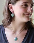 A woman wearing a silver necklace and matching drop earring with a chunky, textured light blue-green stone pendant, shown in a close-up profile shot.