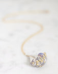 A delicate gold chain necklace with a small, organic-shaped pendant covered in tiny gold rhinestones and a subtle iridescent lavender-blue center, resting on a white marble surface.