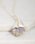 A close-up of a delicate necklace featuring a sailboat-shaped pendant with a silver hull, gold accents, and a translucent iridescent heart-shaped glass center, resting on a light marble surface.