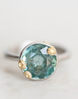 A close-up shot of a ring featuring a round faceted light blue-green gemstone in a silver-toned setting with three small gold-toned accents, resting on a light neutral surface.