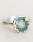 A close-up shot of a ring featuring a round faceted aquamarine gemstone in a bezel setting with textured gold accents on a silver band, set against a soft, light gray background.