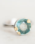 A close-up shot of a ring featuring a faceted light blue-green gemstone in a silver-toned setting with three small gold-toned flower-like accents, on a light neutral background.