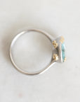 A silver ring with a small oval light blue gemstone and four textured gold accent prongs, photographed on a light neutral surface.