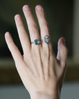 A close-up of a hand wearing a silver ring with a round blue gemstone, with a small geometric tattoo on the middle finger, set against a softly blurred dark background.