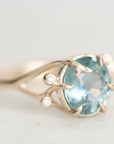 A close-up shot of a gold ring featuring a large oval-cut light blue gemstone, accented by two small round-cut diamonds on the band, set against a soft, light gray background.