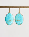 A pair of oval turquoise drop earrings with gold-toned prong settings and hooks, hanging from a gold rod against a light gray background.