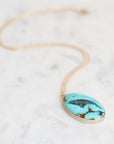 An oval turquoise pendant necklace with a gold-toned bezel and chain, resting on a white marble surface. The pendant features a black bird motif over a vibrant turquoise background with brown matrix details.