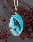 An oval pendant necklace featuring a turquoise stone with a black ink-style illustration of a whale, suspended from a gold chain against a dark gray background with blurred dried red flowers.