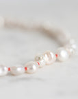 A close-up of a beaded necklace featuring lustrous white baroque pearls separated by thin red string, with a small silver-toned charm, resting on a light marble surface.