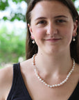 A woman with long brown hair wearing a white freshwater pearl necklace and matching pearl drop earrings, photographed outdoors with a soft green bokeh background.