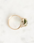 A gold ring with a marquise-cut green gemstone, photographed from above on a light gray marble surface.