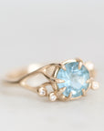 A gold ring featuring a round light blue gemstone in a prong setting, accented with small clear stones along the band, shown on a light gray background.