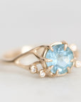 A gold ring featuring a round light blue gemstone in a prong setting, accented with small clear stones along the band, shown on a light gray background.