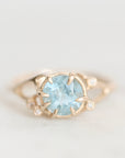 A gold ring featuring a round light blue gemstone in a prong setting, accented with small clear stones on the band, photographed against a soft light gray background.