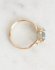 A delicate gold ring with a small light blue gemstone, photographed from above on a light marble surface.