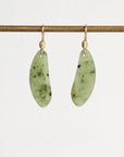 A pair of organic-shaped light green jadeite pendants with dark green inclusions, suspended from gold fishhook ear wires on a gold display bar, against a clean white background.