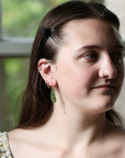 A close-up portrait of a woman with brown hair wearing a small green leaf-shaped dangle earring and multiple hoop earrings, standing in front of a softly blurred window.