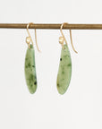 A pair of gold-tone dangle earrings with elongated, polished light green stone pendants containing dark inclusions, hanging from a horizontal gold display rod against a plain white background.