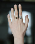 A close-up of a person's hand showing two rings: a gold ring with a small dark green gemstone on the ring finger and a silver-toned geometric ring on the middle finger, against a blurred dark background.