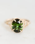 An oval-cut green gemstone ring set in a gold prong setting, shown on a light, neutral background.
