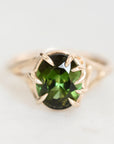 An oval-cut green gemstone ring set in a gold prong setting, shown on a light, neutral background.